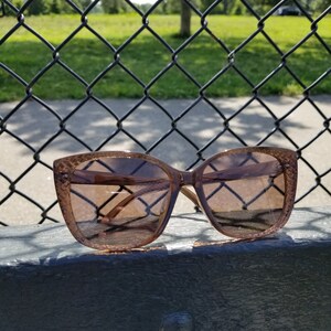 May include: Brown cat-eye sunglasses with a textured frame. The sunglasses are positioned on a dark surface, with a chain-link fence and a park visible in the background. The lenses have a brown gradient tint.
