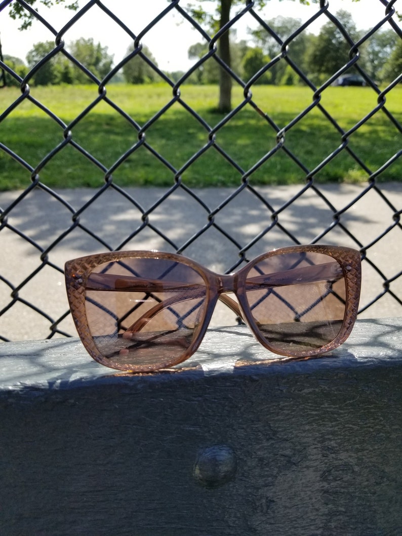 May include: Brown cat-eye sunglasses with a textured frame. The sunglasses are positioned on a dark surface, with a chain-link fence and a park visible in the background. The lenses have a brown gradient tint.