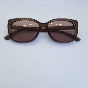 May include: Brown-tinted sunglasses with a textured frame. The lenses are a matching brown colour, and the frame has a subtle, patterned design around the edges. The sunglasses have a classic, slightly oversized shape.