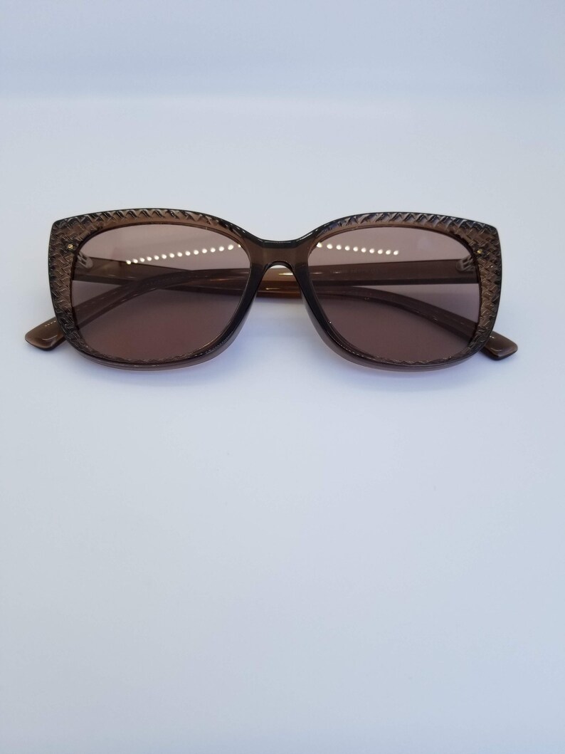 May include: Brown-tinted sunglasses with a textured frame. The lenses are a matching brown colour, and the frame has a subtle, patterned design around the edges. The sunglasses have a classic, slightly oversized shape.