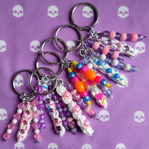 May include: Five colorful keychains with white, pink, purple, blue, and orange beads and charms. Each keychain features a white swirl with sprinkles and a heart charm.
