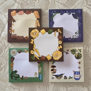 May include: Five square notepads with decorative borders. Themes include food like pasta and cheese, and nature scenes with a frog, bird, and mushrooms. Each notepad has a white center for writing.