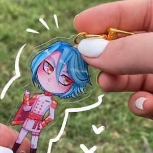May include: A clear acrylic keychain featuring a cartoon character with blue hair, red and white clothing, and a gold chain. The character is standing with a hand on their hip and has a surprised expression.