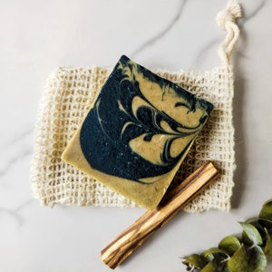 Puede incluir: Una barra de jabón artesanal con un patrón de remolino negro y amarillo. El jabón está sentado en una bolsa de malla de fibra natural con un palo de madera al lado.