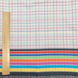 May include: White fabric with a multi-colored plaid pattern. The fabric has a border of black, red, blue, green, orange, pink, turquoise, and yellow stripes.