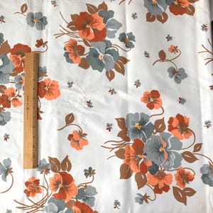 May include: White fabric with a repeating floral pattern. The flowers are in shades of orange, gray, and brown, with green accents. A wooden ruler is placed vertically on the left side of the fabric, measuring approximately 12 inches.