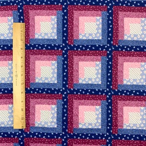 May include: Close-up of a fabric with a repeating pattern of squares. Each square features concentric rectangles in shades of pink, blue, and burgundy, with a floral center. A wooden ruler is placed vertically along the left side, showing inch measurements.