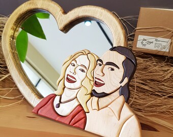 Op de afbeelding: Een hartvormige houten spiegel met een gesneden houten stel erin. De spiegel is gemaakt van lichtbruin hout en het stel is gemaakt van verschillende tinten hout. Het stel glimlacht en kijkt naar elkaar.