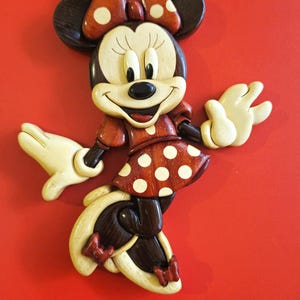 Könnte beinhalten: Eine Holzfigur von Minnie Maus, in Rot, Weiß und Braun bemalt. Sie trägt ein rotes Kleid mit weißen Punkten und eine rote Schleife mit weißen Punkten. Sie lächelt und hat ihre Arme ausgestreckt.