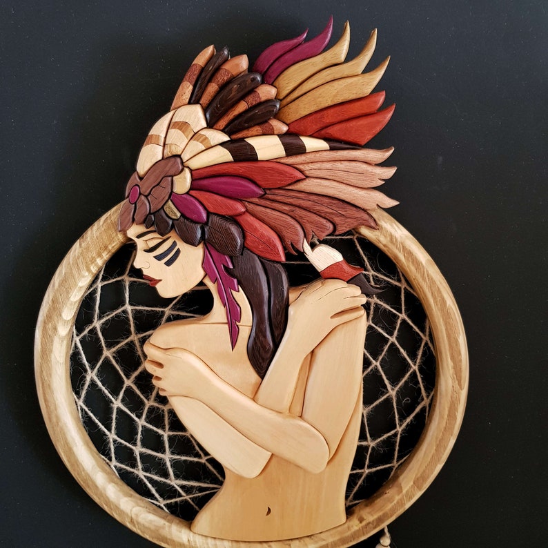 May include: A wooden sculpture of a woman with a feathered headdress, holding her arms around herself. The sculpture is set against a brown background and is surrounded by a dreamcatcher.