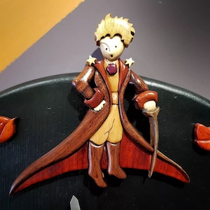 Peut inclure: Une figurine en bois du Petit Prince, un personnage du livre d'Antoine de Saint-Exupéry. La figurine est peinte dans des tons de brun, rouge et jaune. Le Petit Prince porte un manteau rouge et tient une épée. La figurine est montée sur un fond vert foncé.