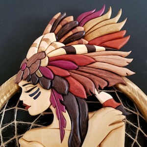 Może przedstawiać: Drewniana rzeźba przedstawiająca indiańską kobietę w ozdobnym nakryciu głowy. Twarz kobiety jest pomalowana czarnymi i białymi liniami. Rzeźba jest umieszczona na tle łapacza snów.