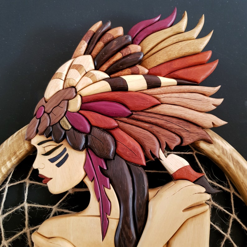 May include: Intricately carved wooden sculpture of a Native American woman wearing a headdress with feathers in shades of red, brown, and gold. The woman is holding a dreamcatcher with a woven web.