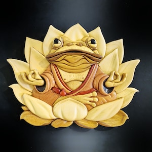 May include: A wooden sculpture of a frog sitting in a lotus flower position. The frog is wearing a red robe and has a serene expression on its face. The sculpture is made of light and dark wood, giving it a natural and rustic look.