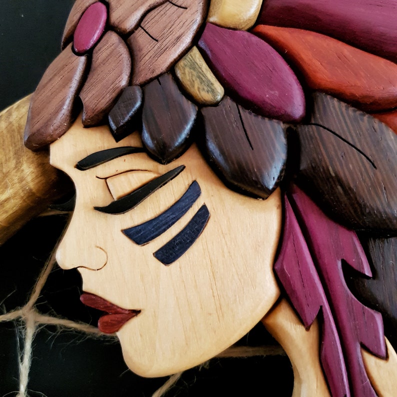 May include: A wooden carving of a woman's face with a feathered headdress. The woman has dark eyes, a red mouth, and blue and black markings on her face. The headdress is made of red, brown, and purple feathers.