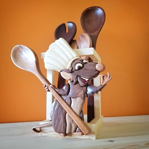Puede incluir: Un soporte de utensilios de cocina de madera con un chef de rata tallado que lleva un gorro de chef blanco. La rata sostiene una cuchara de madera. El soporte tiene ranuras para cucharas, espátulas y otros utensilios de cocina.