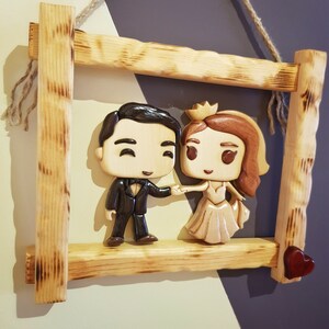 Op de afbeelding: Een houten frame met een gesneden houten stel dat elkaars hand vasthoudt. Het stel is gestileerd met grote hoofden en kleine lichamen. Het frame hangt aan een touwtje en heeft een klein rood hartje in de rechter benedenhoek.