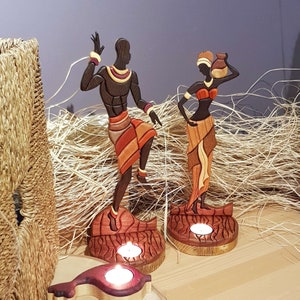 May include: Wooden candle holders featuring carved figures of a man and woman in traditional African attire. The figures are painted in shades of brown, red, and yellow. The candle holders are designed to hold tea lights.