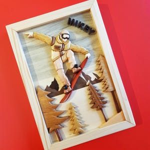 May include: Wooden framed artwork featuring a snowboarder carving down a mountain slope. The snowboarder is wearing a yellow and tan outfit with goggles. The word "MIKEY" is above the snowboarder. The artwork includes wooden trees and mountains.