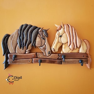 May include: Wooden wall-mounted key holder featuring two horse heads facing each other. The horses are carved from various shades of wood, with dark brown and tan manes. The key holder has four metal hooks and is mounted on a wooden fence.