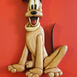 Może przedstawiać: Drewniana figurka Plutona, kreskowego psa z Disneya, siedzącego z wystawionym językiem. Figurka wykonana jest z jasnobrązowego drewna z ciemnobrązowymi akcentami na uszach, nosie i oczach.