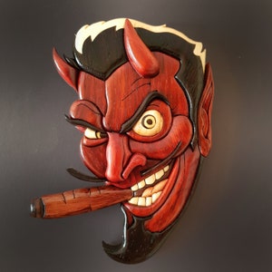 以下が含まれることがあります： いたずらっぽい笑顔でシガーをくわえた赤い悪魔の木彫り。悪魔は角ととがったあごひげ、そして細部まで作り込まれた顔をしています。