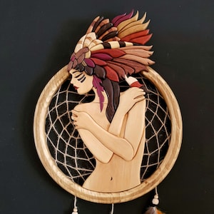 May include: A wooden dreamcatcher with a carved woman's face and a colorful feathered headdress. The dreamcatcher is made of wood and has a brown rope web.