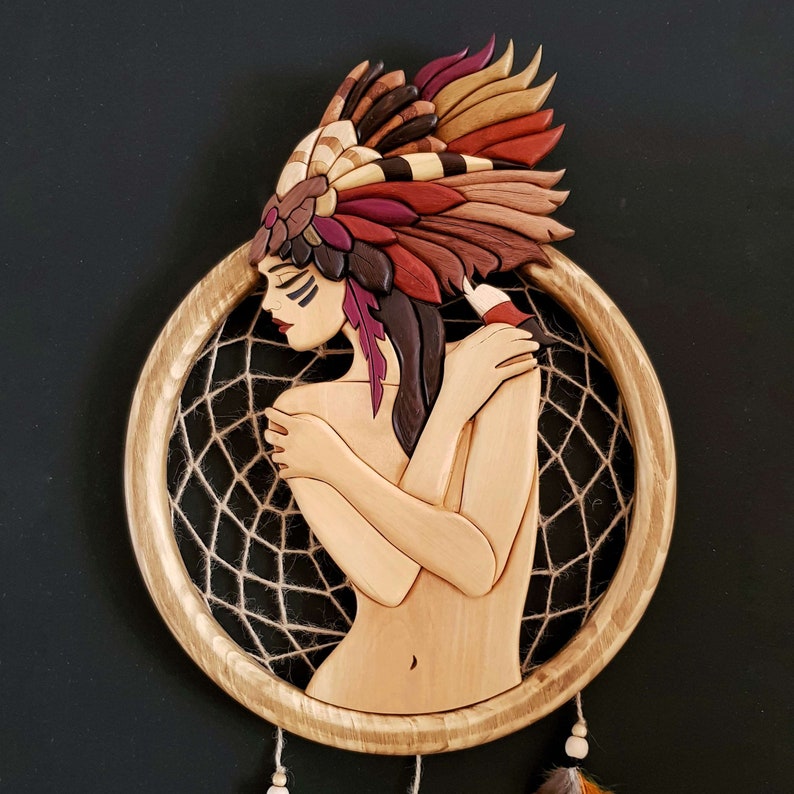 May include: A wooden dreamcatcher with a carved woman's face and a colorful feathered headdress. The dreamcatcher is made of wood and has a brown rope web.