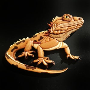 Może przedstawiać: Szczegółowa drewniana rzeźba iguany, wykonana z różnych odcieni brązu i beżu. Iguana ma teksturowane ciało z ciemnobrązowymi plamami i kolczastym grzebieniem. Rzeźba jest umieszczona na czarnym tle, podkreślając kunszt wykonania.