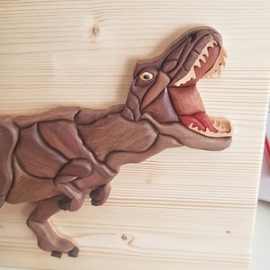 Peut inclure: Une sculpture en bois d'un dinosaure Tyrannosaurus Rex avec la bouche ouverte, montrant ses dents. La sculpture est faite de différentes nuances de bois brun et est montée sur un fond en bois clair.