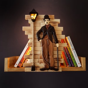 May include: Wooden wall shelf with a carved figure of Charlie Chaplin holding a cane, standing next to a lit street lamp. The shelf has a row of books on the right side.