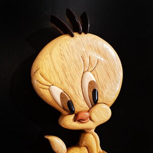 Puede incluir: Una talla de madera de Piol&iacute;n, un p&aacute;jaro de dibujos animados amarillo con ojos negros, un pico negro y una cresta negra. La talla es detallada y realista, con una textura de veta de madera.
