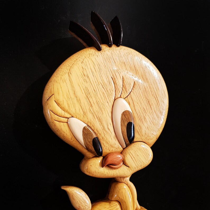 Puede incluir: Una talla de madera de Piol&iacute;n, un p&aacute;jaro de dibujos animados amarillo con ojos negros, un pico negro y una cresta negra. La talla es detallada y realista, con una textura de veta de madera.