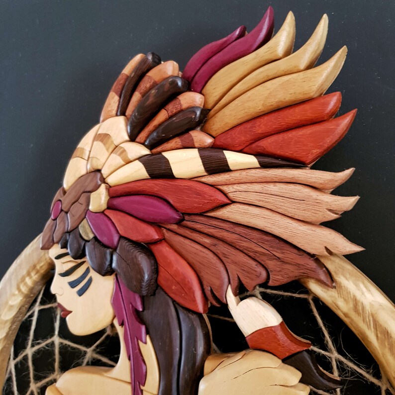 May include: A wooden sculpture of a Native American woman wearing a headdress with intricate details. The headdress is made of various shades of brown, red, and orange wood. The woman's face is carved with delicate features and she is holding a dreamcatcher.