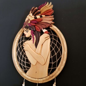May include: A wooden dreamcatcher with a carved female figure wearing a colorful headdress. The figure is holding her arms around herself and the dreamcatcher is made of brown wood with a brown and white string web.