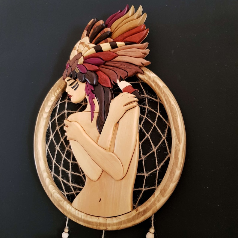 May include: A wooden dreamcatcher with a carved female figure wearing a colorful headdress. The figure is holding her arms around herself and the dreamcatcher is made of brown wood with a brown and white string web.