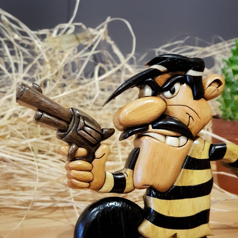 May include: A wooden figurine of a cartoon character with a black and yellow striped shirt, holding a brown wooden gun. The character has a large nose, a black mustache, and a scowling expression.