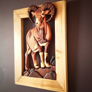 Puede incluir: Una escultura de pared de madera de una cabra mont&eacute;s marr&oacute;n y blanca con grandes cuernos. La escultura est&aacute; enmarcada en un marco de madera natural.