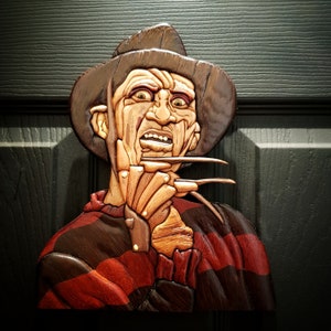 Pode incluir: Uma escultura de madeira de Freddy Krueger, o personagem de terror fictício da série de filmes A Hora do Pesadelo. Ele está usando um chapéu fedora marrom, um suéter listrado vermelho e marrom e tem uma expressão ameaçadora em seu rosto. Sua icônica luva com garras é visível.