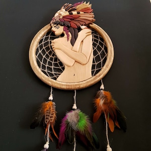 May include: A wooden dreamcatcher with a carved female figure wearing a headdress. The dreamcatcher has a brown wooden frame with a woven net and three feathers hanging from the bottom.