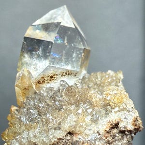 Op de afbeelding: Een helder kwartskristal met een puntige top, met interne regenbogen. Het kristal staat op een cluster van kleinere, lichtgekleurde kristallen en een ruwe, bruine basis. Natuurlijke en geologische uitstraling.