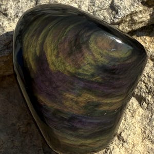 Puede incluir: Una piedra de obsidiana pulida de forma ovalada. La piedra presenta un patr&oacute;n arremolinado de colores iridiscentes, incluyendo p&uacute;rpura, verde y dorado. La superficie es lisa y reflectante, con un acabado brillante. La piedra descansa sobre una superficie rocosa rugosa y texturizada.