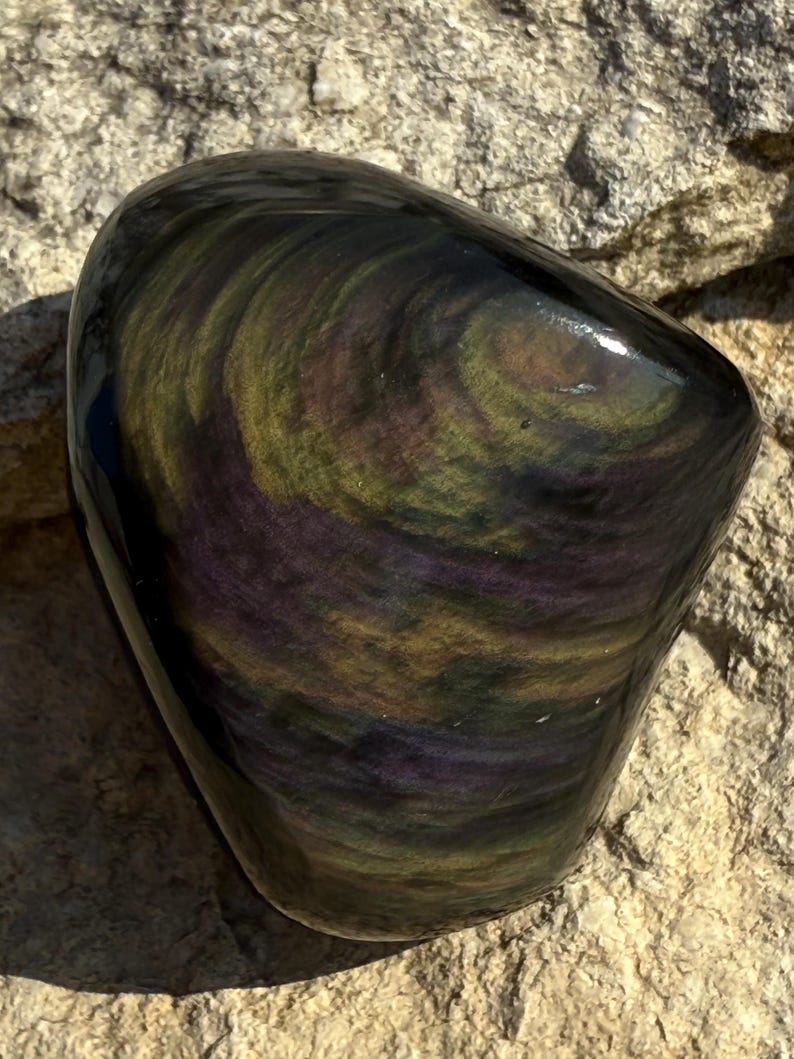 Puede incluir: Una piedra de obsidiana pulida de forma ovalada. La piedra presenta un patr&oacute;n arremolinado de colores iridiscentes, incluyendo p&uacute;rpura, verde y dorado. La superficie es lisa y reflectante, con un acabado brillante. La piedra descansa sobre una superficie rocosa rugosa y texturizada.