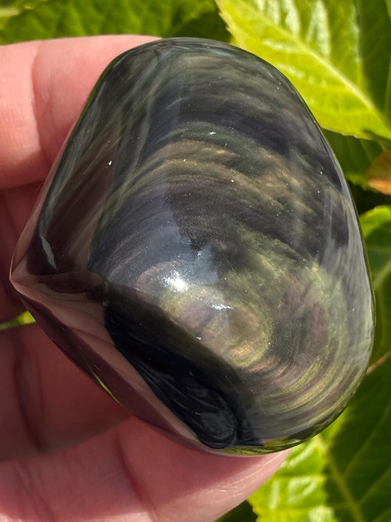 Puede incluir: Una piedra de obsidiana pulida con patrones arremolinados de negro, verde y dorado. La forma lisa y redondeada de la piedra se sostiene en una mano, con follaje verde en el fondo. La piedra tiene una superficie brillante y reflectante.