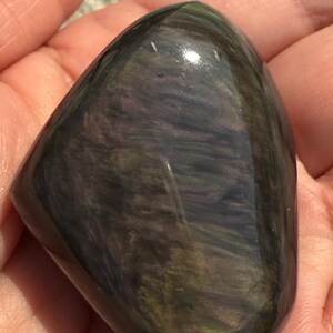 Puede incluir: Una piedra de obsidiana pulida e iridiscente. La piedra tiene una forma lisa y redondeada, mostrando un patr&oacute;n arremolinado de colores verde oscuro, p&uacute;rpura y negro. La superficie refleja la luz, resaltando la belleza natural de la piedra.