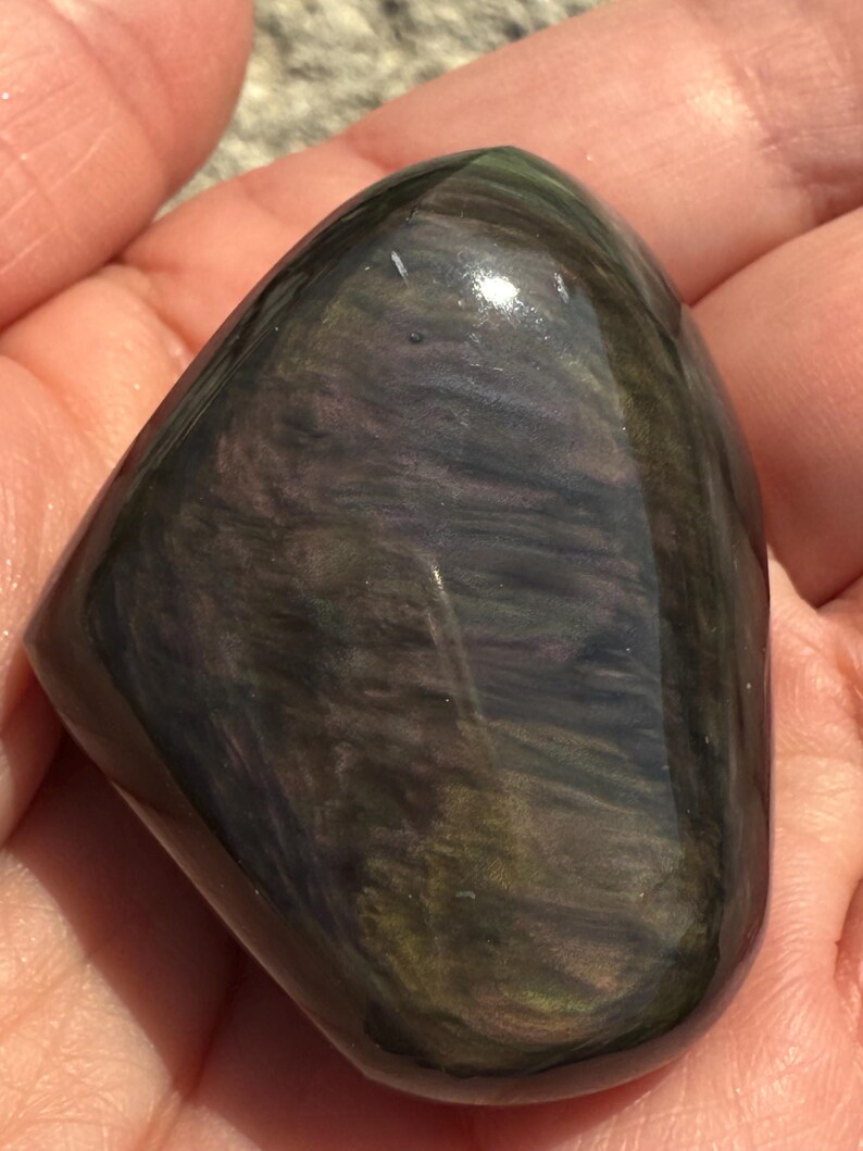 Puede incluir: Una piedra de obsidiana pulida e iridiscente. La piedra tiene una forma lisa y redondeada, mostrando un patr&oacute;n arremolinado de colores verde oscuro, p&uacute;rpura y negro. La superficie refleja la luz, resaltando la belleza natural de la piedra.