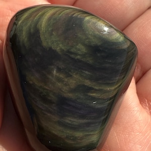 Puede incluir: Una piedra de obsidiana pulida de forma ovalada. La piedra presenta patrones arremolinados de verde oscuro, dorado y negro. La superficie es lisa y reflectante, con un acabado brillante. La piedra se sostiene en una mano, mostrando su belleza natural.