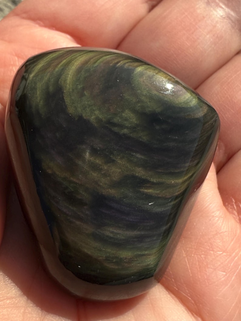 Puede incluir: Una piedra de obsidiana pulida de forma ovalada. La piedra presenta patrones arremolinados de verde oscuro, dorado y negro. La superficie es lisa y reflectante, con un acabado brillante. La piedra se sostiene en una mano, mostrando su belleza natural.