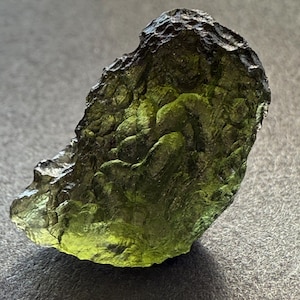 Puede incluir: Una gema natural rugosa de color verde y negro, con una forma única e irregular.