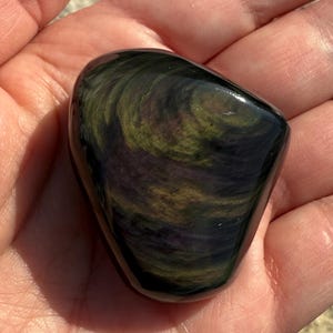 Puede incluir: Una piedra de obsidiana pulida con patrones arremolinados de verde, dorado y p&uacute;rpura. La piedra es lisa y redondeada, sostenida en una mano abierta, y refleja la luz, mostrando sus colores y texturas naturales.